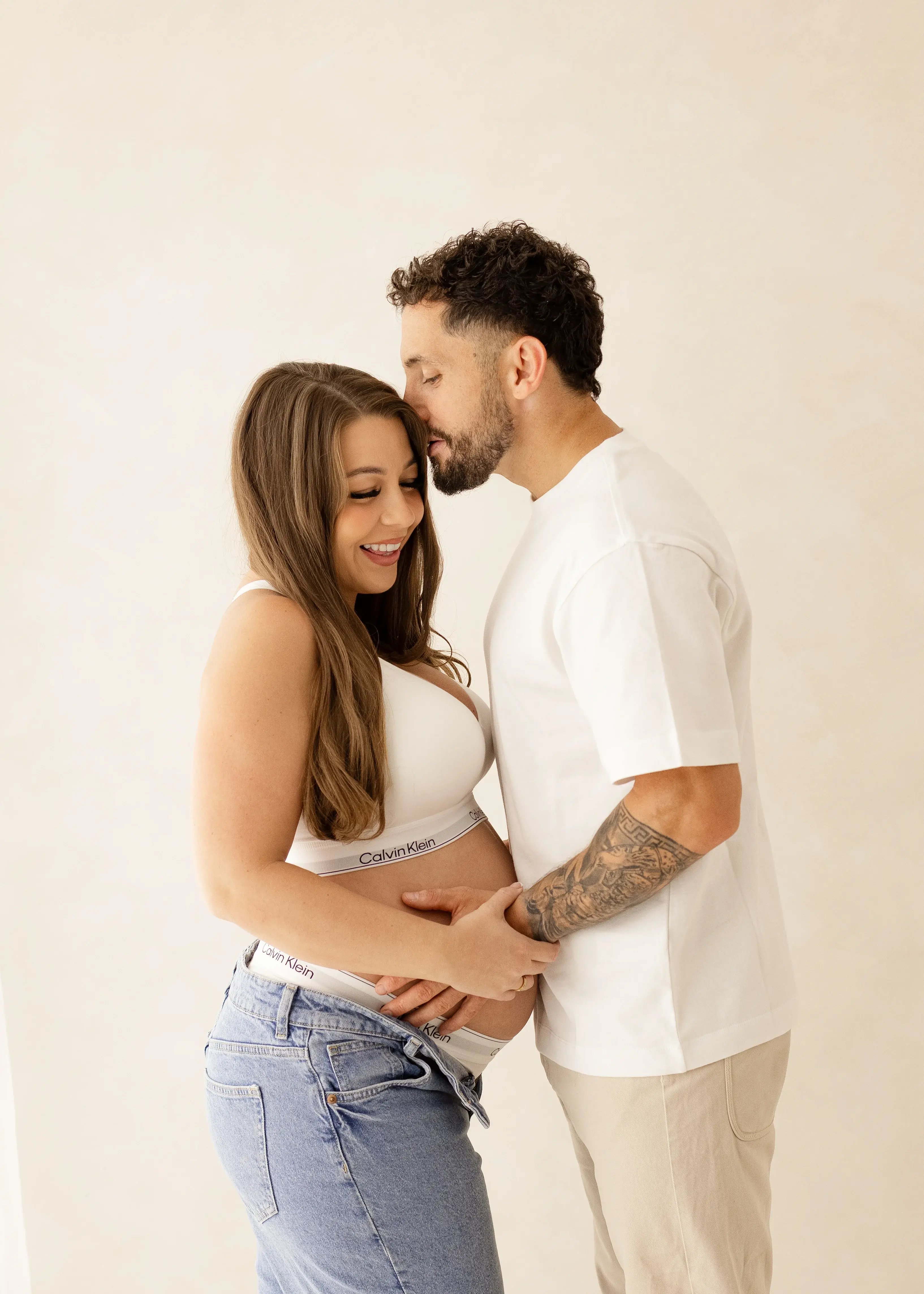 Expectant couple smiling and holding the baby bump, with the partner kissing her forehead during a soft, natural maternity photoshoot in Kent.