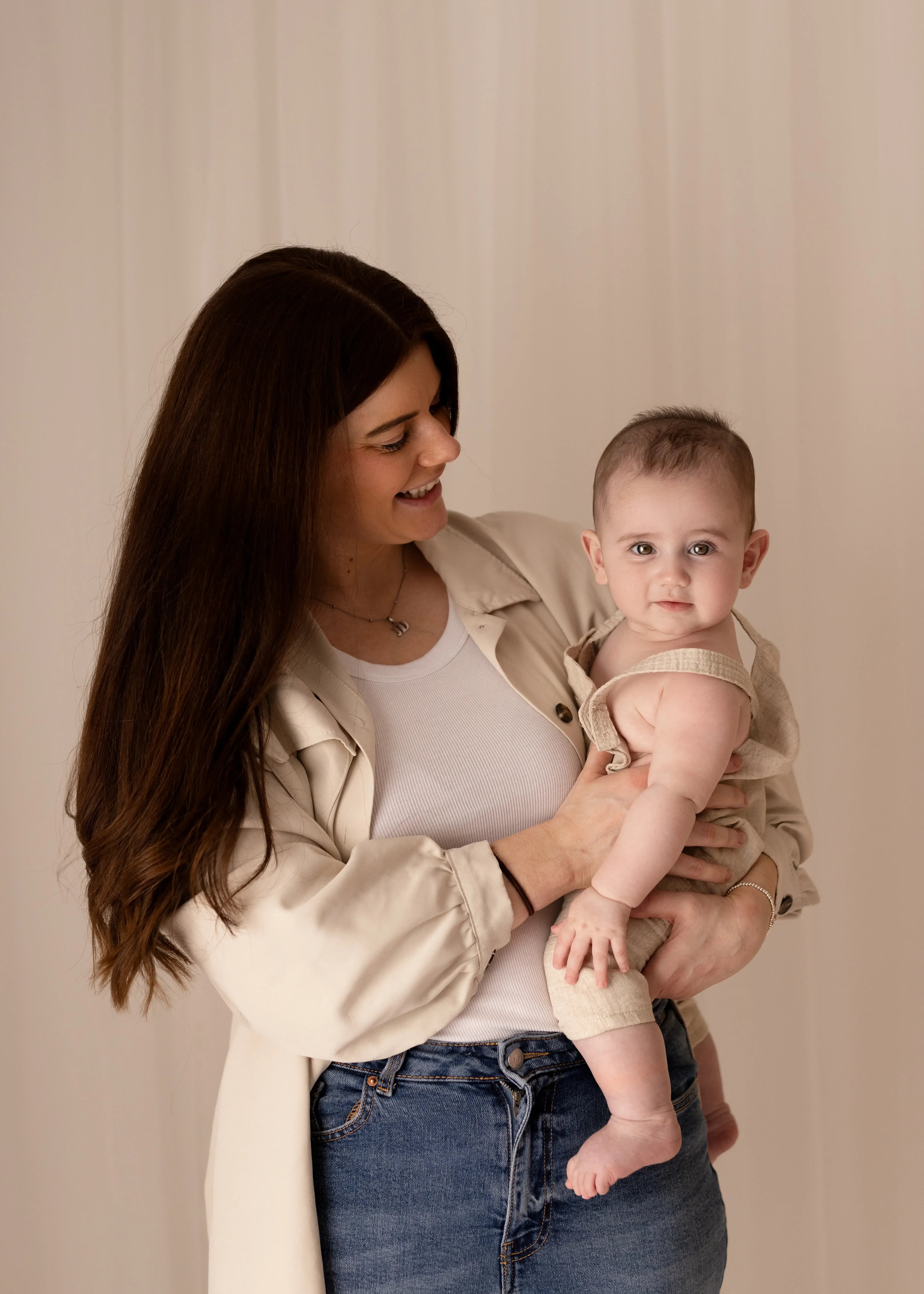 Smiling mother with dark hair wearing a neutral shirt and jeans, holding her baby in light beige dungarees. The baby gazes at the camera, conveying a calm moment during a relaxed family photography session.