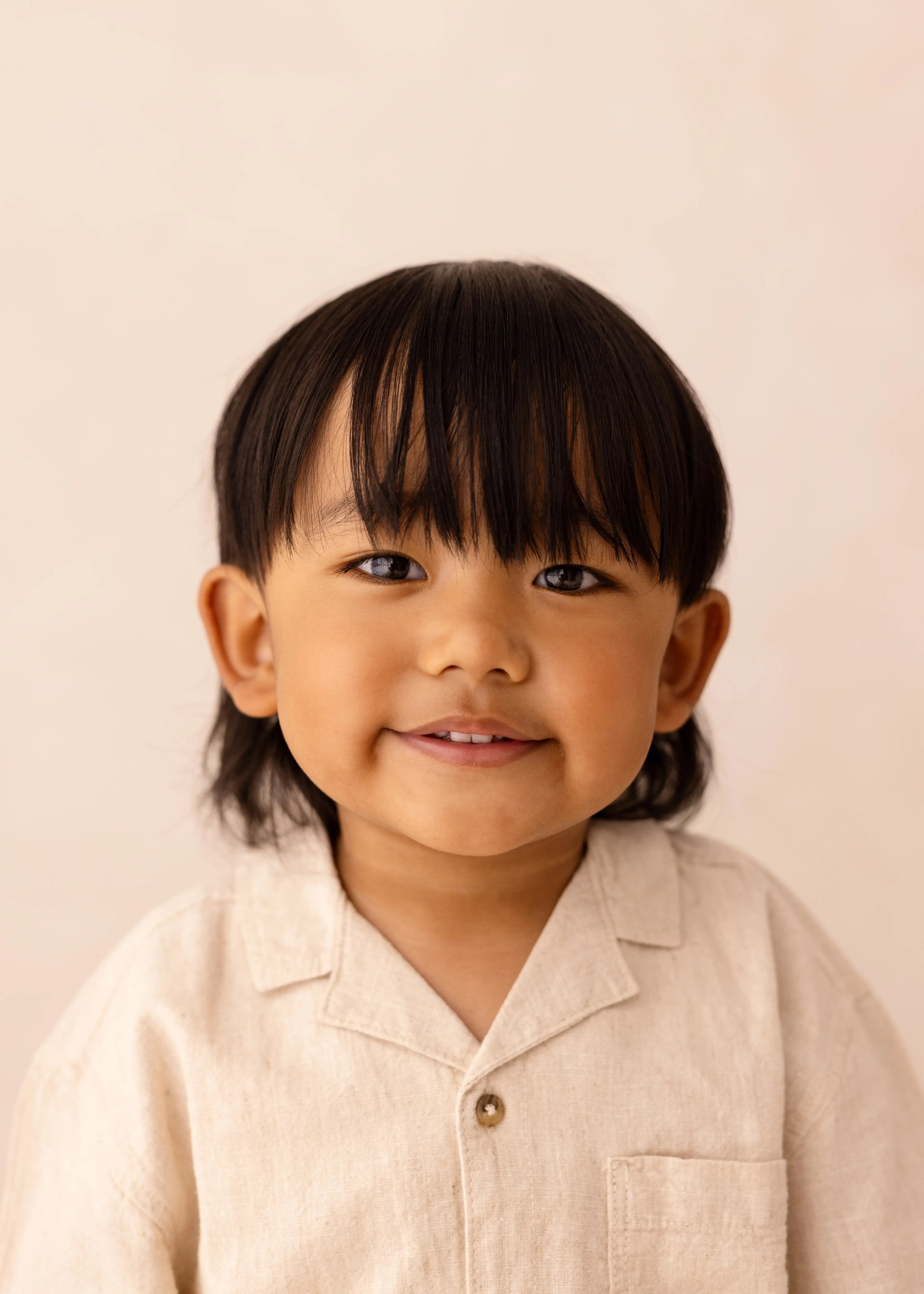 Smiling young child with long dark hair wearing a beige shirt in a minimalist studio portrait session.