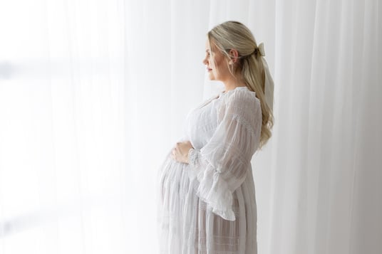 Mum-to-be looking out of a window in a white maternity gown for maternity photoshoot