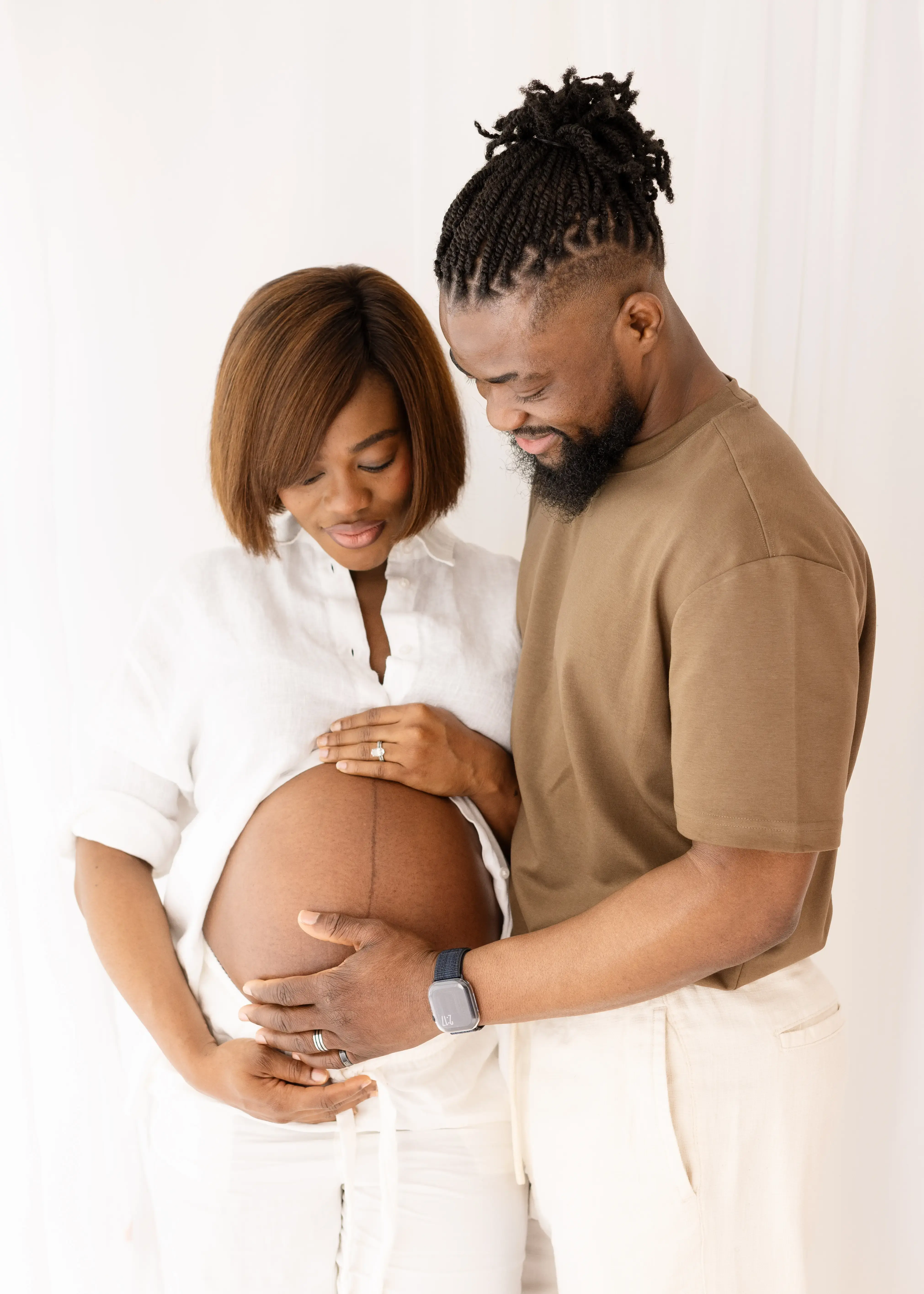 Close-up of an expectant couple gently cradling a baby bump together, both looking down tenderly with soft smiles, dressed in white and earth tones against a light studio backdrop.